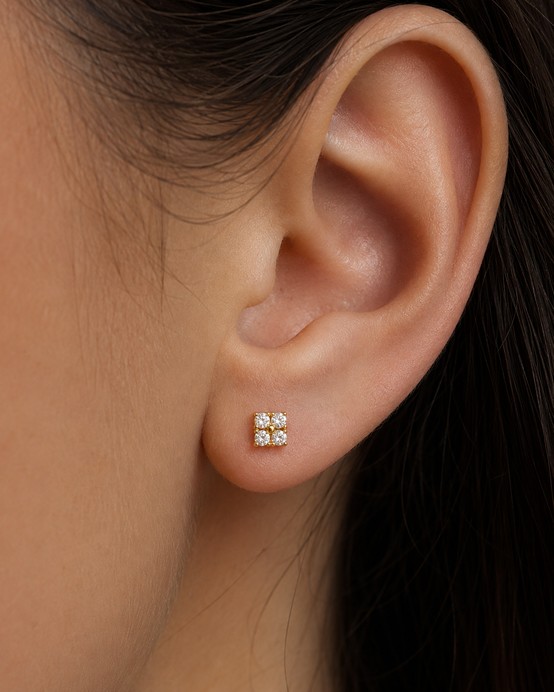 A close-up of a person's ear wearing a small square stud earring with four clear gemstones in a gold setting.