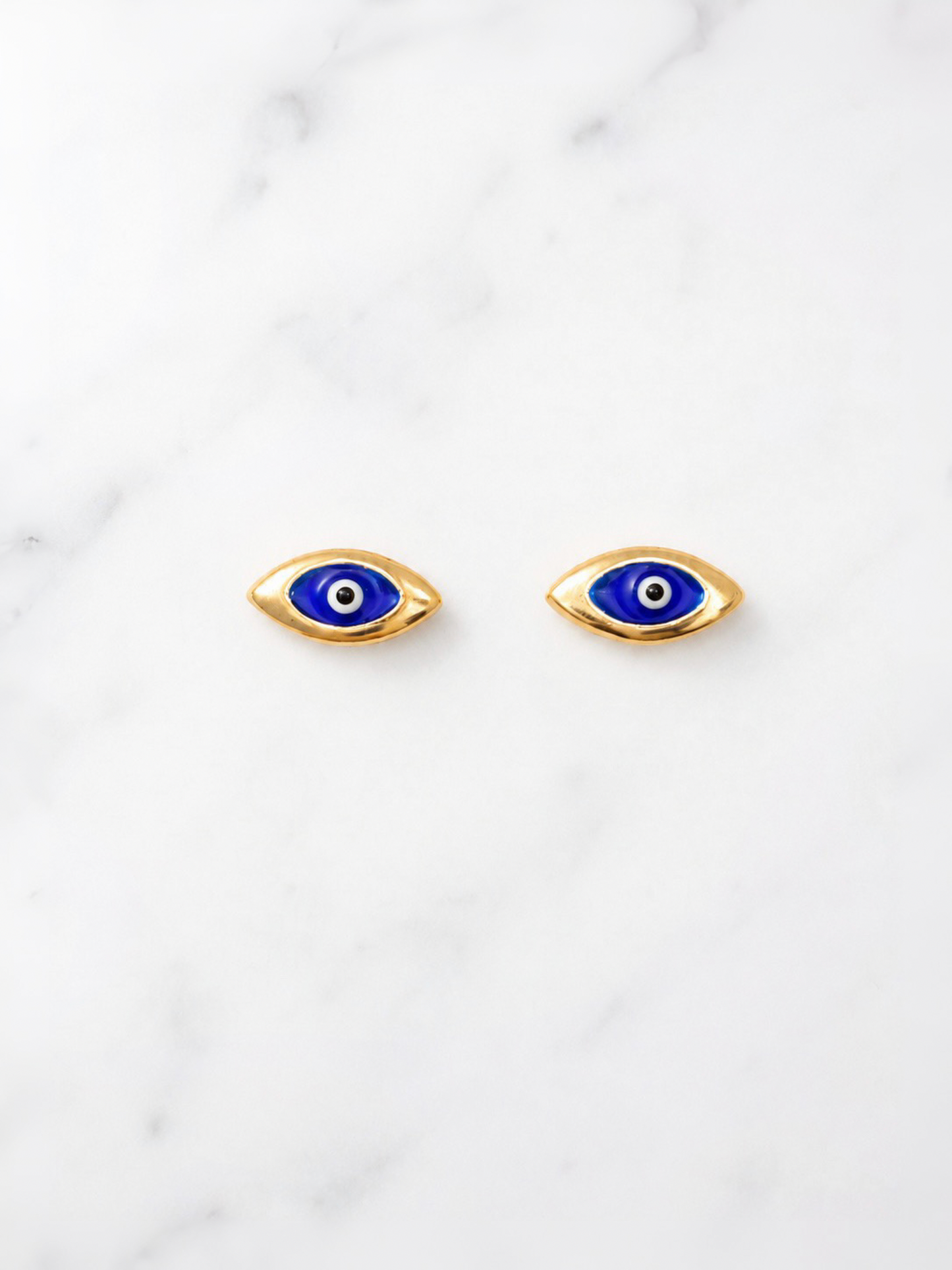 A pair of gold-toned evil eye stud earrings with blue, white, and black details, displayed on a white marble surface.
