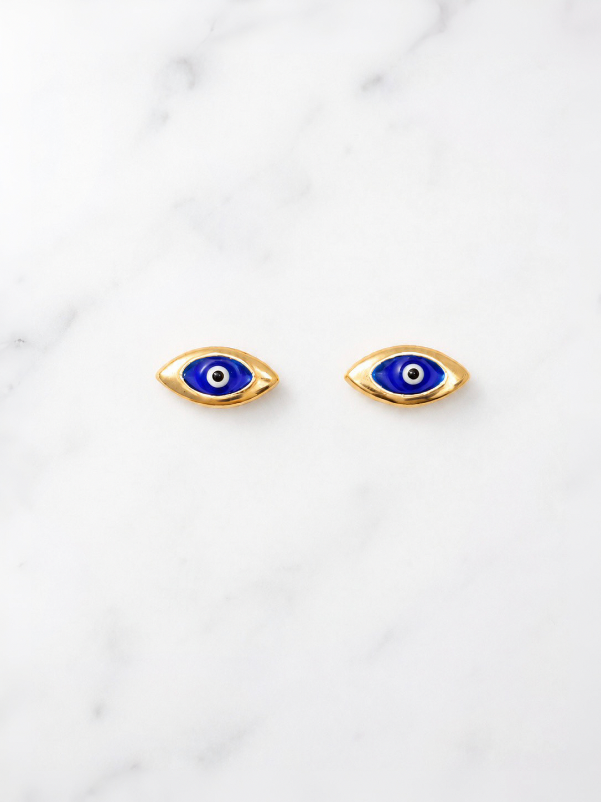 A pair of gold-toned evil eye stud earrings with blue, white, and black details, displayed on a white marble surface.