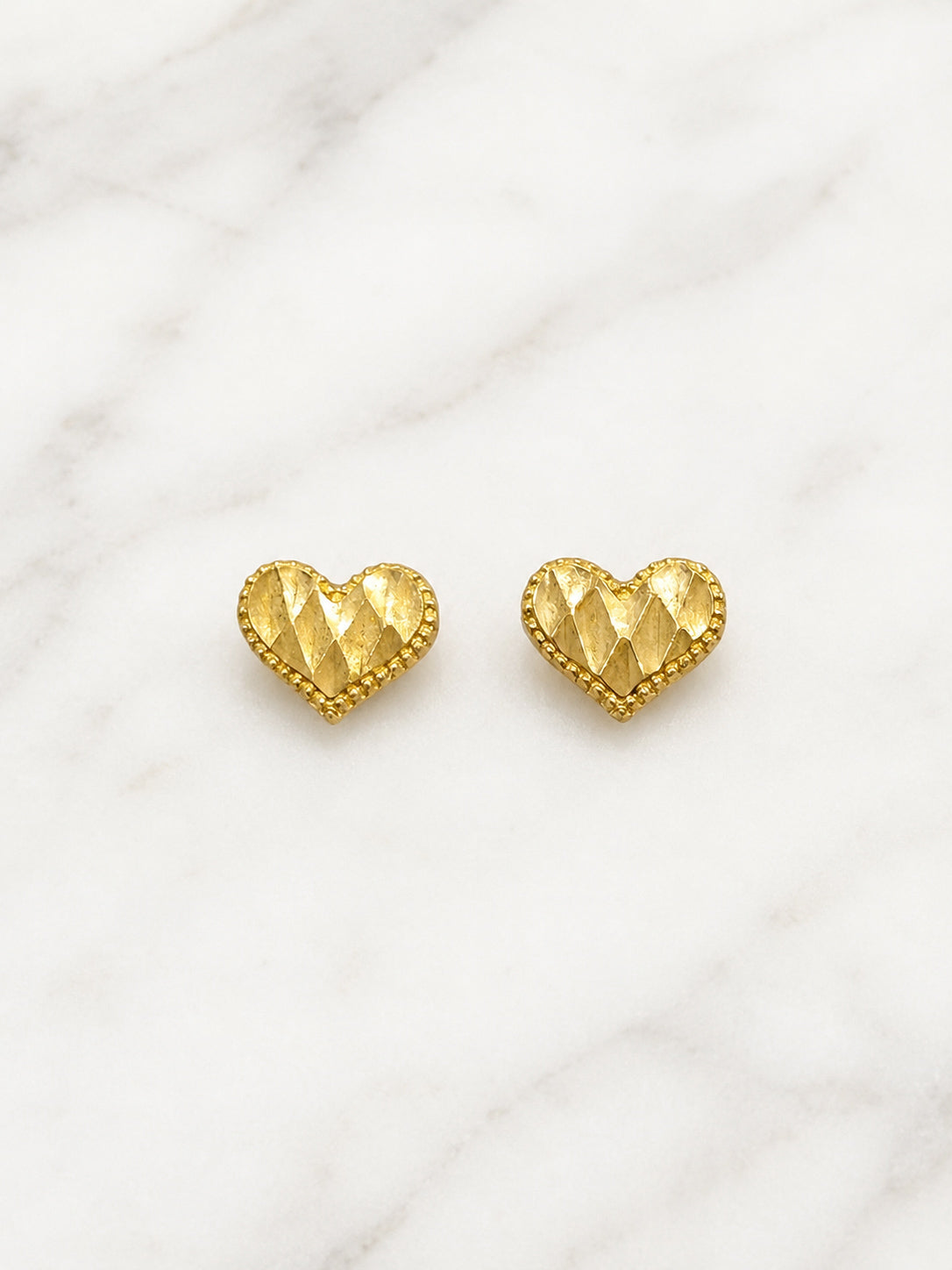 A pair of gold heart-shaped stud earrings with a textured, faceted diamond pattern and a beaded border, displayed on a white marble background.