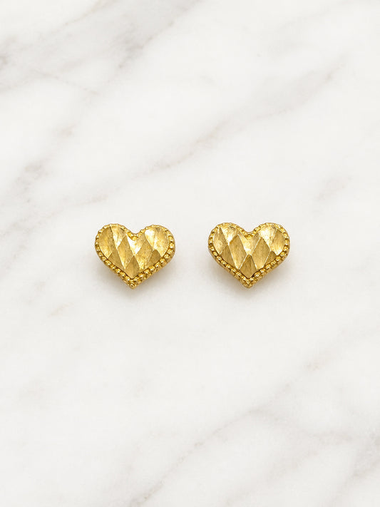 A pair of gold heart-shaped stud earrings with a textured, faceted diamond pattern and a beaded border, displayed on a white marble background.