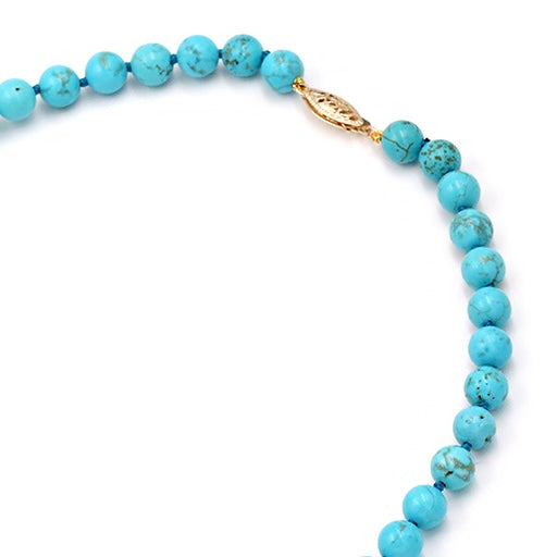 A beaded necklace made of round turquoise-blue beads with natural marbling, featuring a decorative gold-toned clasp, shown on a plain white background.