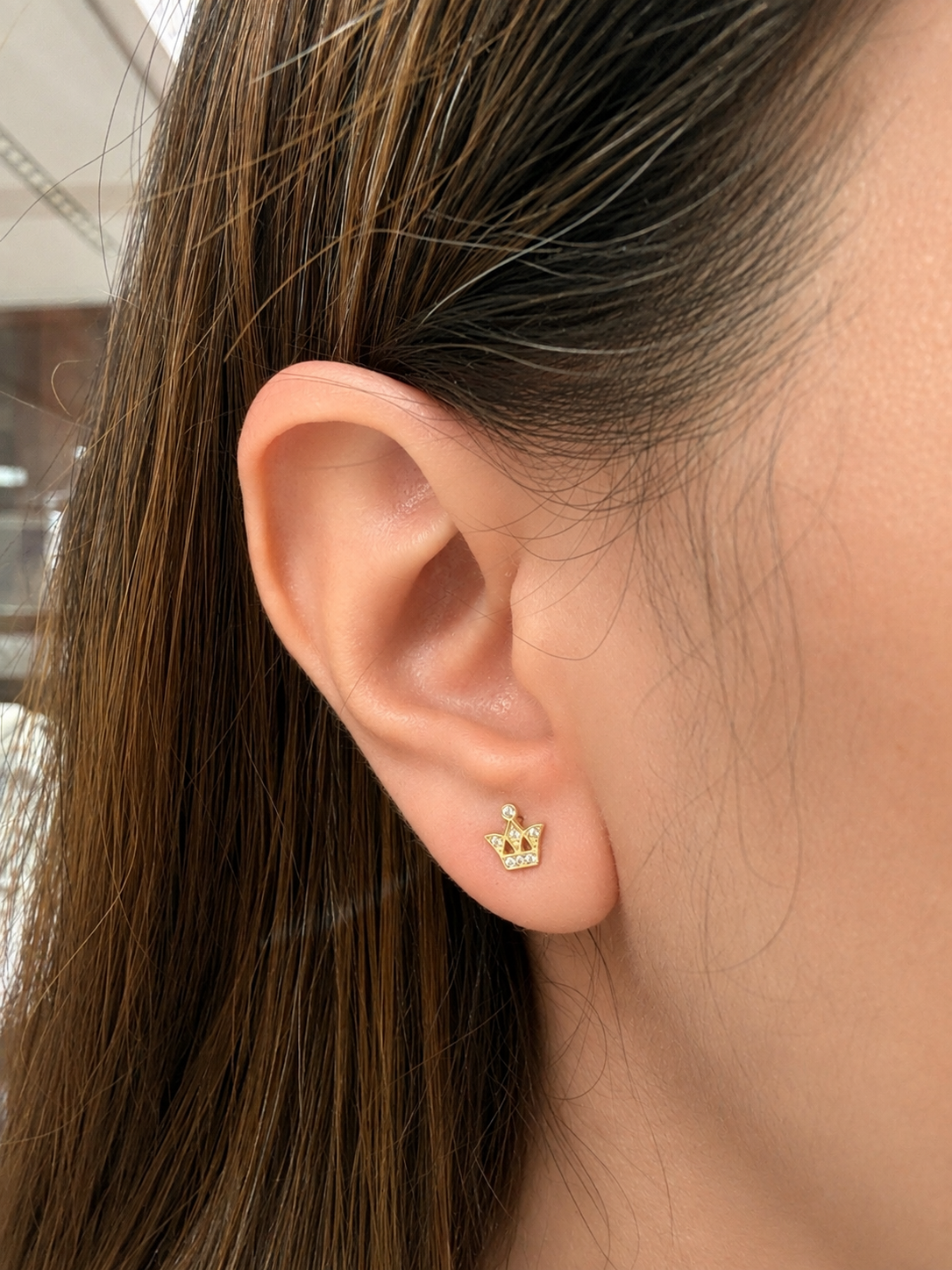 A close-up of a person's ear wearing a small gold crown stud earring with tiny clear stones.
