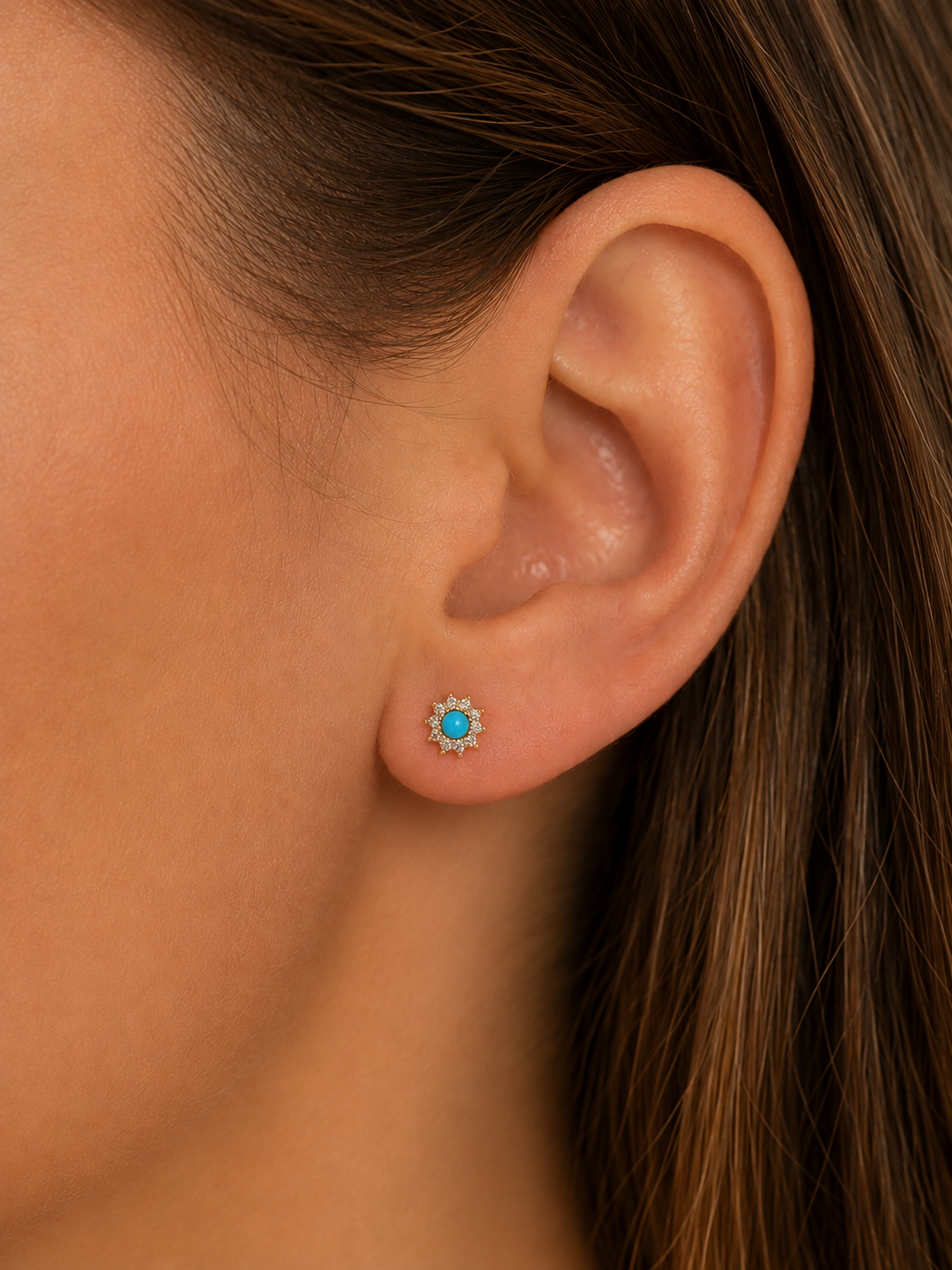 A close-up of a model's ear wearing a small floral stud earring with a bright turquoise blue center and a halo of tiny clear stones.
