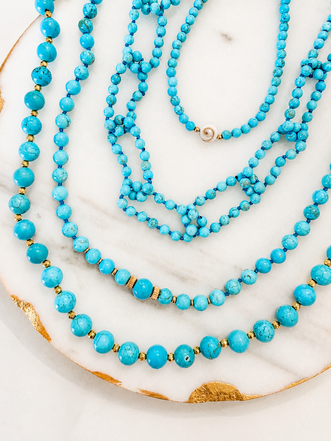 A set of layered turquoise bead necklaces with small gold spacer beads, arranged on a white marble tray with gold leaf accents.