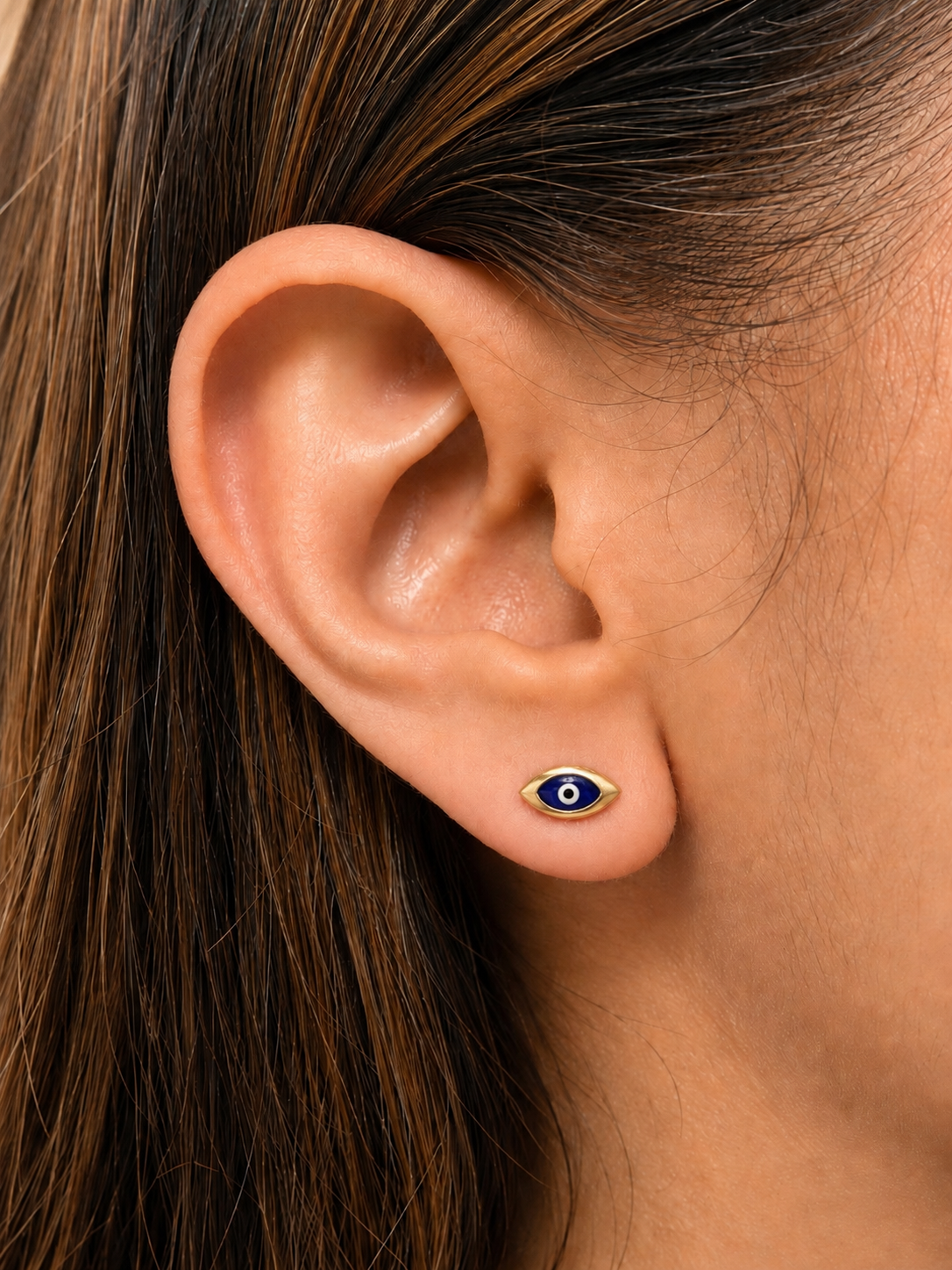 A close-up of a person's ear wearing a small evil eye stud earring with blue, white, and gold details.