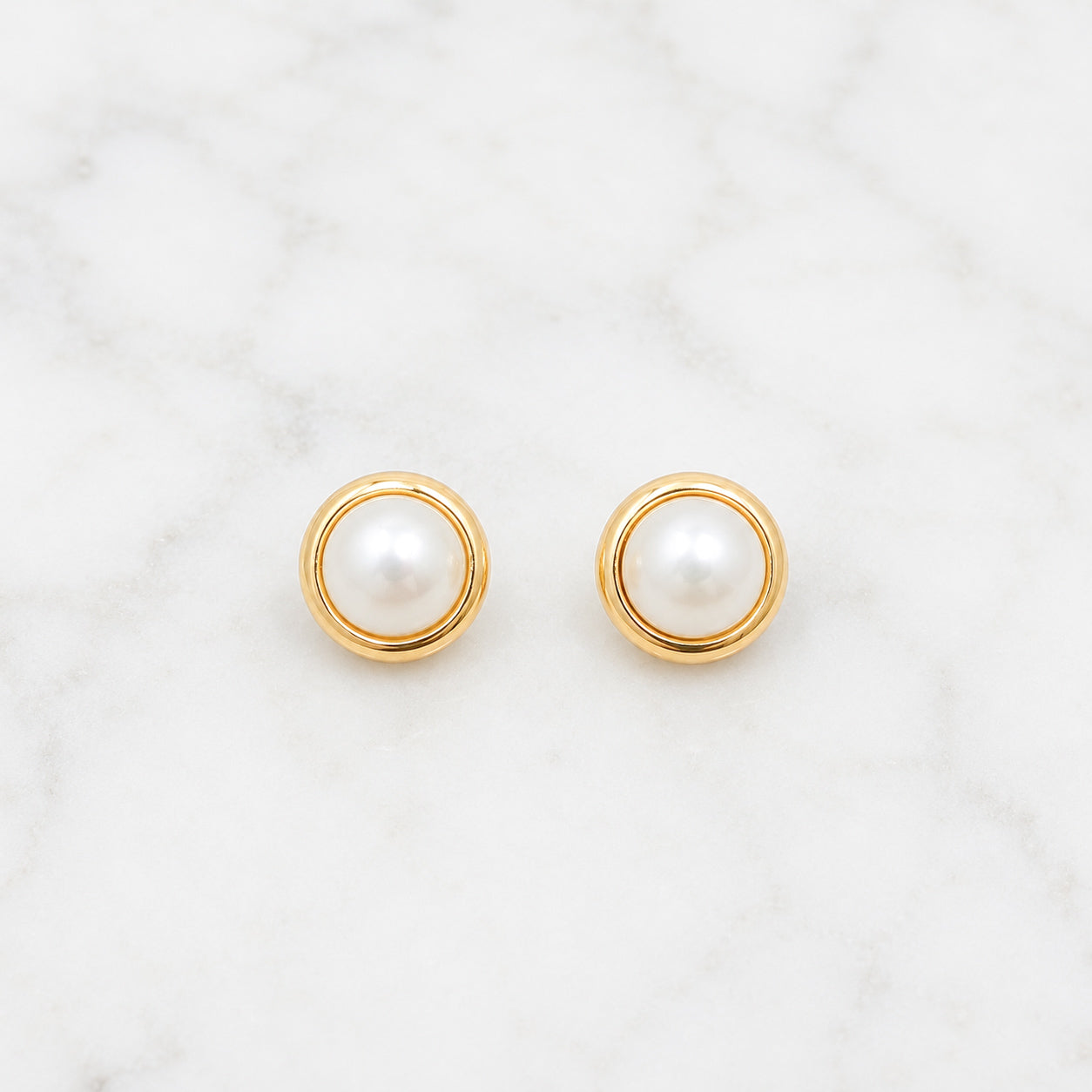 A pair of gold circular stud earrings with white pearl centers, displayed on a white marble surface.