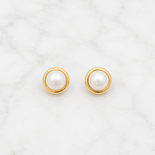 A pair of gold circular stud earrings with white pearl centers, displayed on a white marble surface.