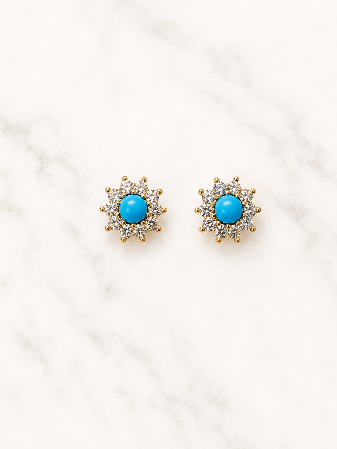 A pair of gold-toned stud earrings with a central round turquoise blue stone surrounded by small clear stones, displayed on a white marble surface.