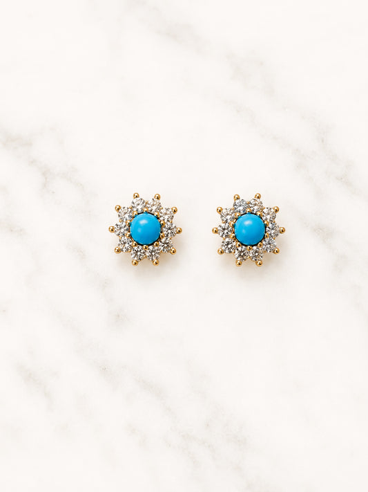 A pair of gold-toned stud earrings with a central round turquoise blue stone surrounded by small clear stones, displayed on a white marble surface.
