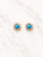 A pair of gold-toned stud earrings with a central round turquoise blue stone surrounded by small clear stones, displayed on a white marble surface.