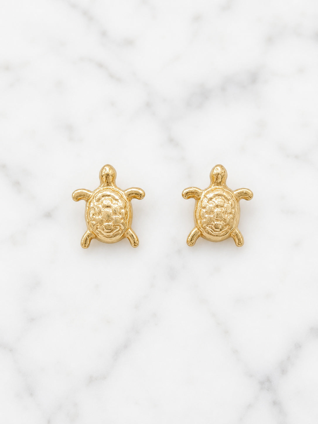 A pair of small gold turtle stud earrings displayed on a white marble surface.