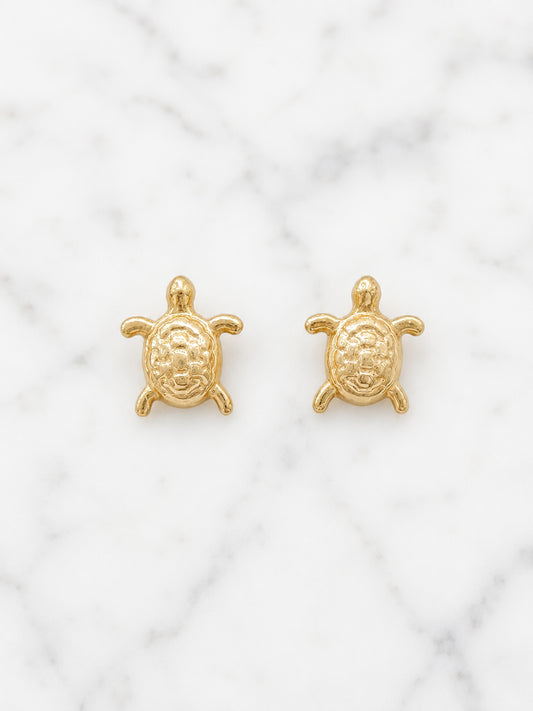 A pair of small gold turtle stud earrings displayed on a white marble surface.