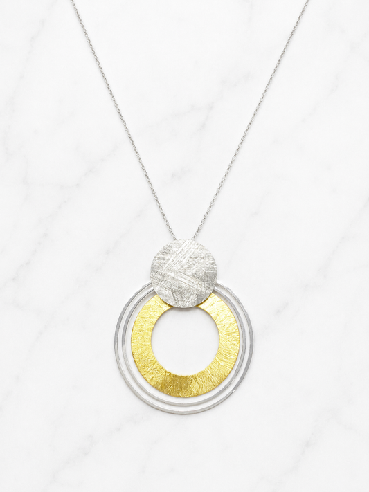 A modern pendant necklace featuring concentric silver rings with a textured gold center, topped with a textured silver disc, suspended from a delicate silver chain on a white marble background.