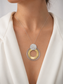A close-up of a model wearing a modern layered necklace with concentric gold and silver rings and a textured silver disc pendant, styled with a white blazer.