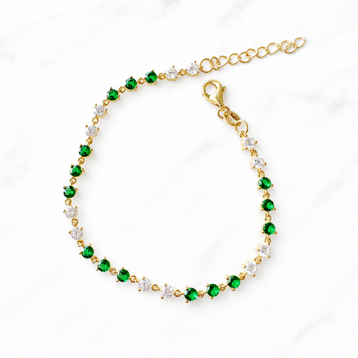 A gold tennis bracelet featuring alternating round-cut emerald green stones and clear cubic zirconia stones, displayed on a white marble background.
