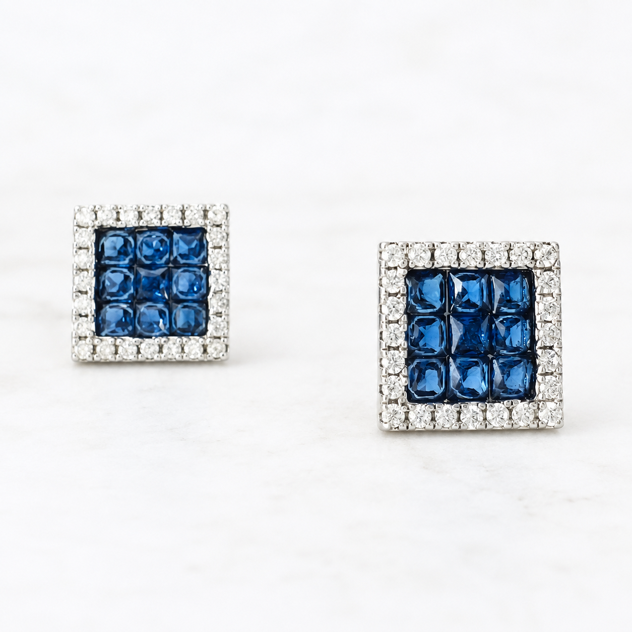 A pair of square stud earrings featuring a central grid of deep blue faceted stones surrounded by a border of small clear stones, set in a silver-toned metal on a white marble background.