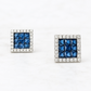 A pair of square stud earrings featuring a central grid of deep blue faceted stones surrounded by a border of small clear stones, set in a silver-toned metal on a white marble background.