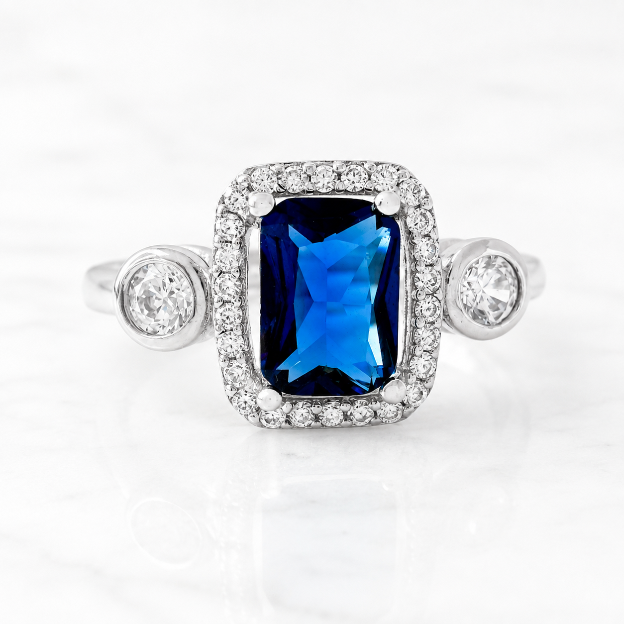 A silver-toned halo ring featuring a rectangular emerald-cut blue sapphire center stone and round clear accent stones on the shoulders, shown on a white marble surface.