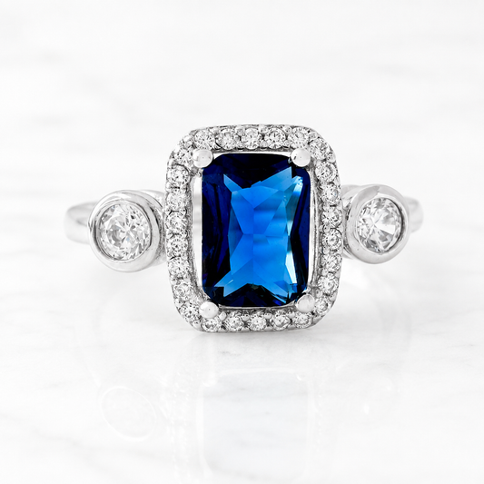 A silver-toned halo ring featuring a rectangular emerald-cut blue sapphire center stone and round clear accent stones on the shoulders, shown on a white marble surface.