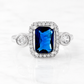 A silver-toned halo ring featuring a rectangular emerald-cut blue sapphire center stone and round clear accent stones on the shoulders, shown on a white marble surface.