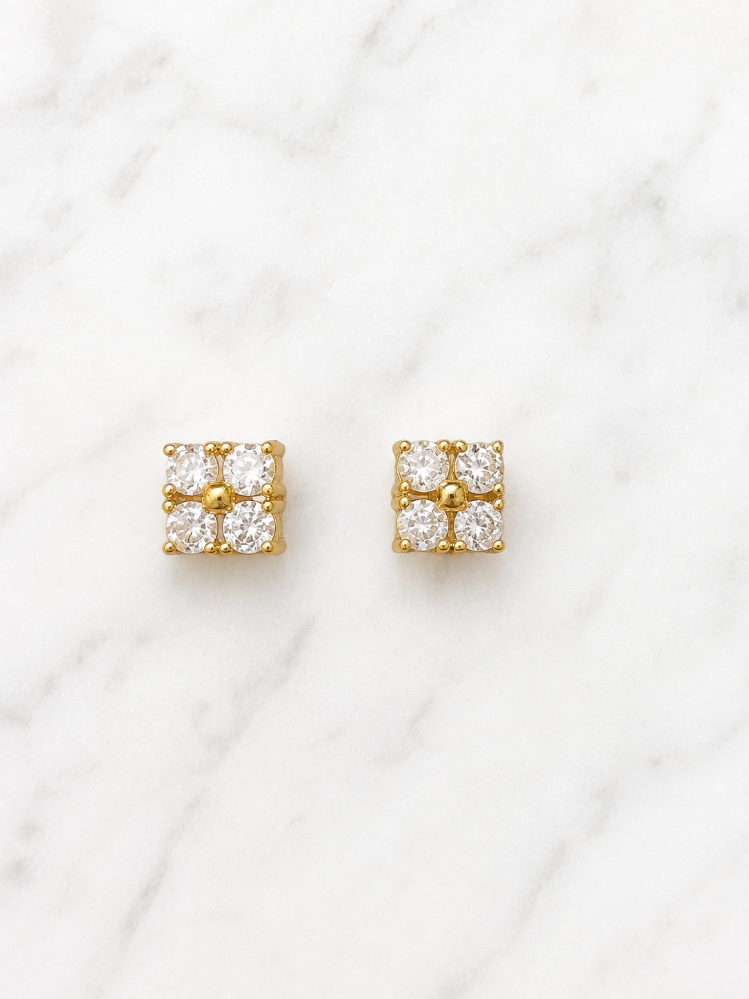 A pair of gold square stud earrings, each set with four clear round-cut gemstones in a four-petal cluster arrangement, displayed on a white marble surface.