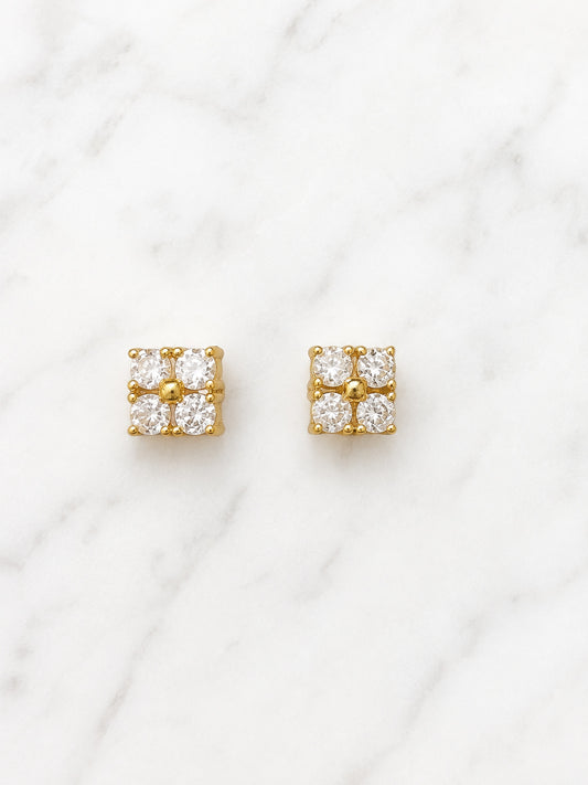 A pair of gold square stud earrings, each set with four clear round-cut gemstones in a four-petal cluster arrangement, displayed on a white marble surface.