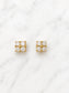 A pair of gold square stud earrings, each set with four clear round-cut gemstones in a four-petal cluster arrangement, displayed on a white marble surface.
