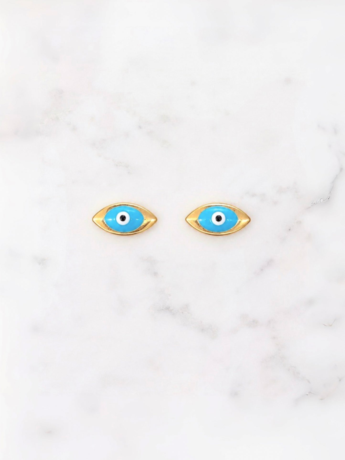 A pair of gold-tone evil eye stud earrings with bright blue enamel, white centers, and black pupils, displayed on a white marble surface.