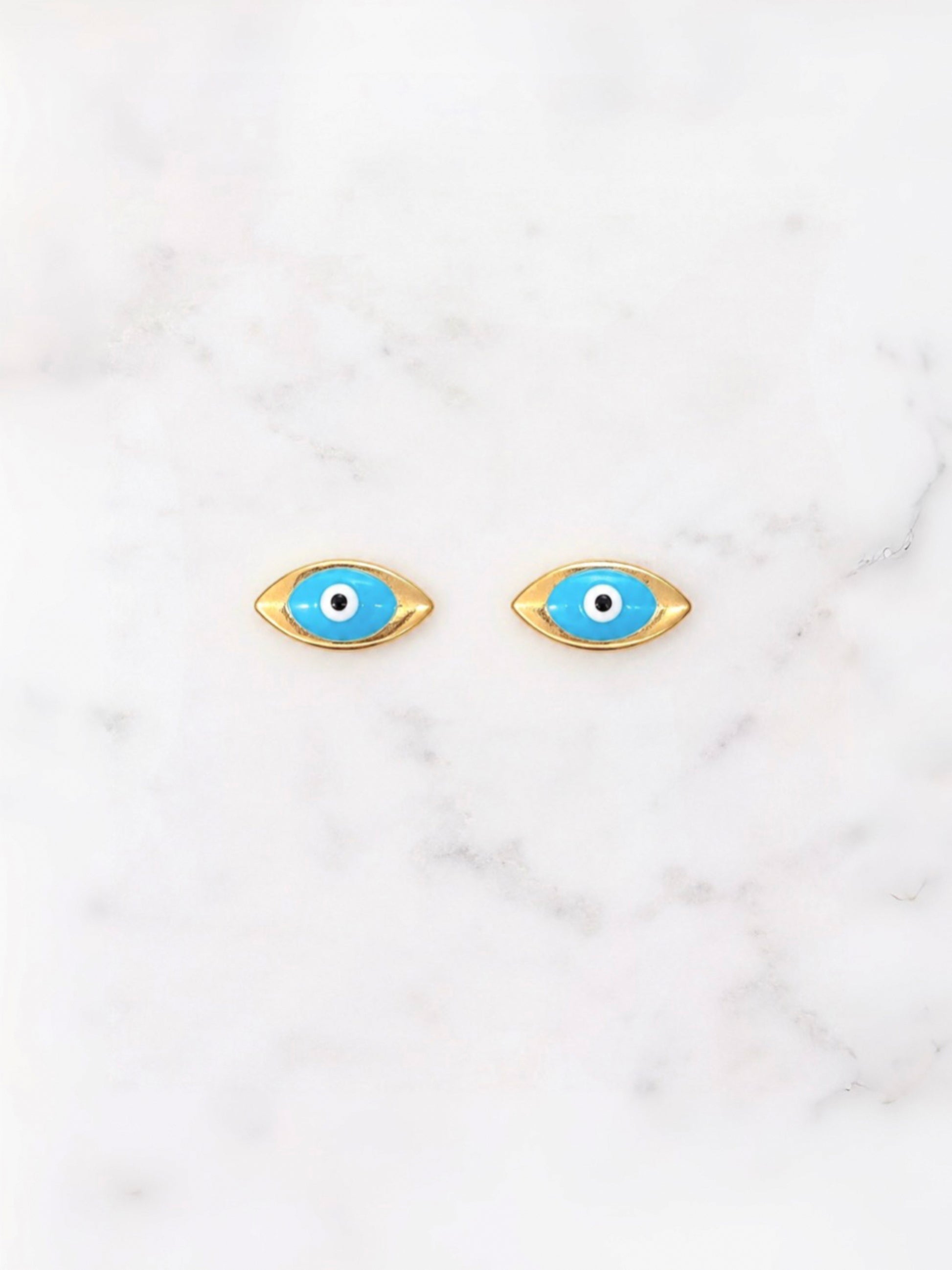 A pair of gold-tone evil eye stud earrings with bright blue enamel, white centers, and black pupils, displayed on a white marble surface.