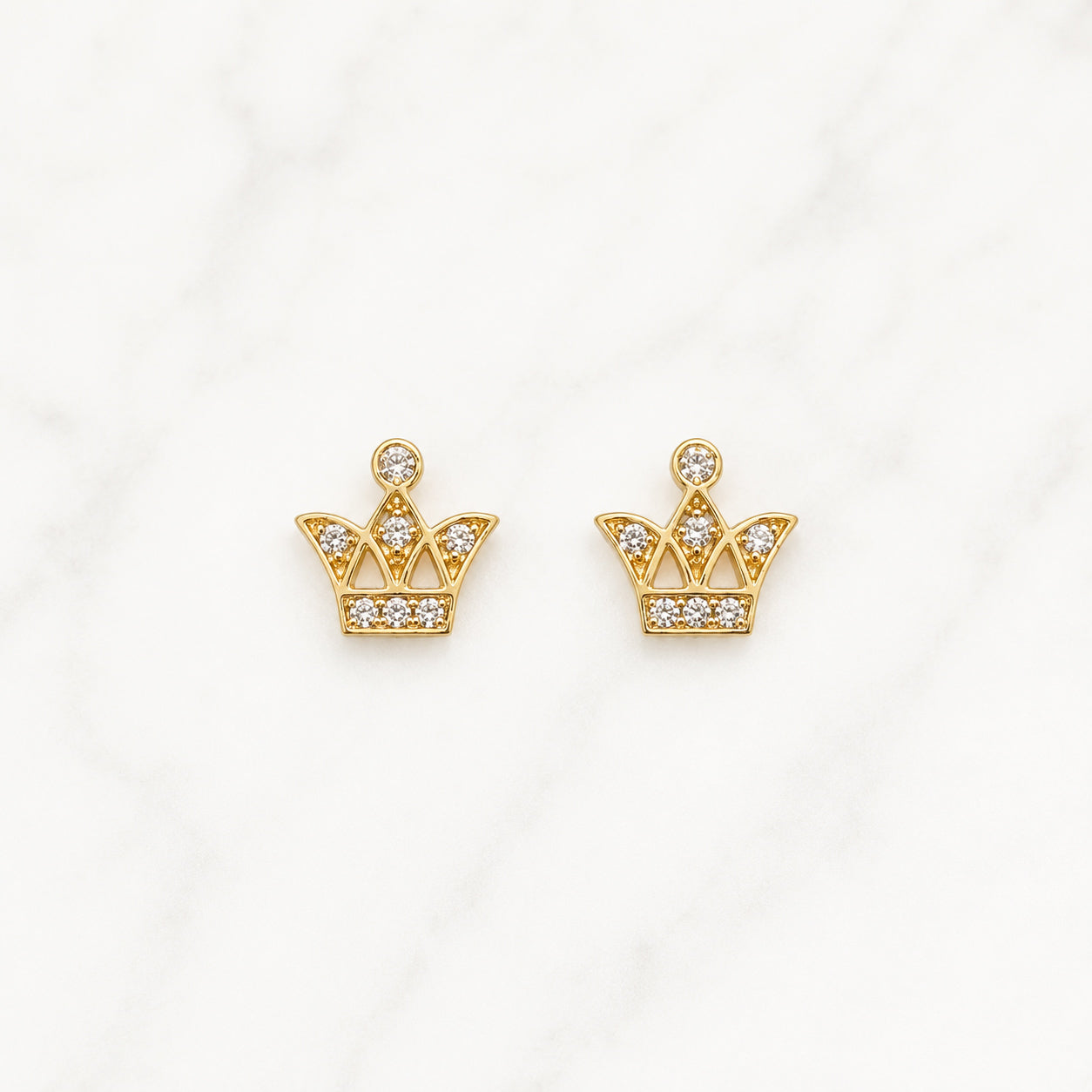 A pair of gold crown-shaped stud earrings set with small clear gemstones, displayed on a white marble surface.