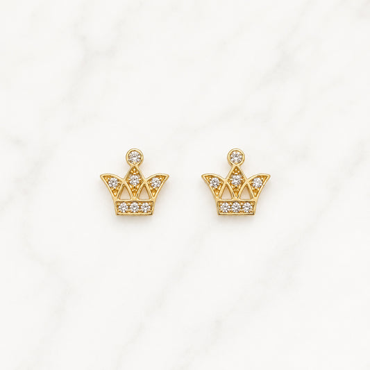 A pair of gold crown-shaped stud earrings set with small clear gemstones, displayed on a white marble surface.