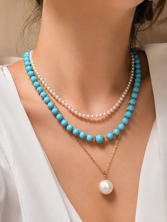 A close-up of a model wearing a layered necklace set. The set includes a double-strand turquoise and gold beaded necklace, a single-strand white pearl necklace, and a gold chain with a single white pearl pendant.