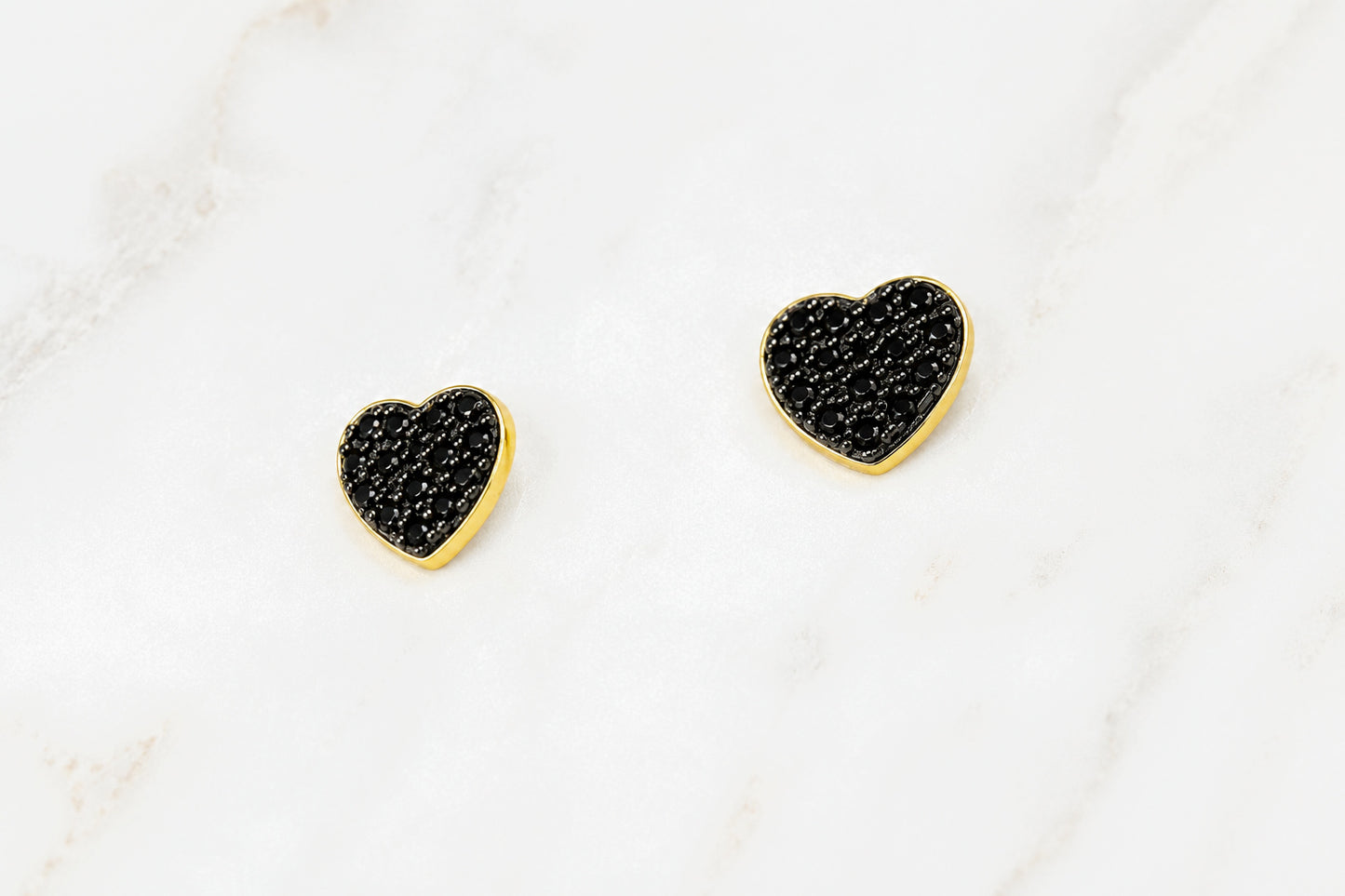 A pair of small heart-shaped stud earrings with textured black pavé centers and polished gold borders, displayed on a white marble surface.