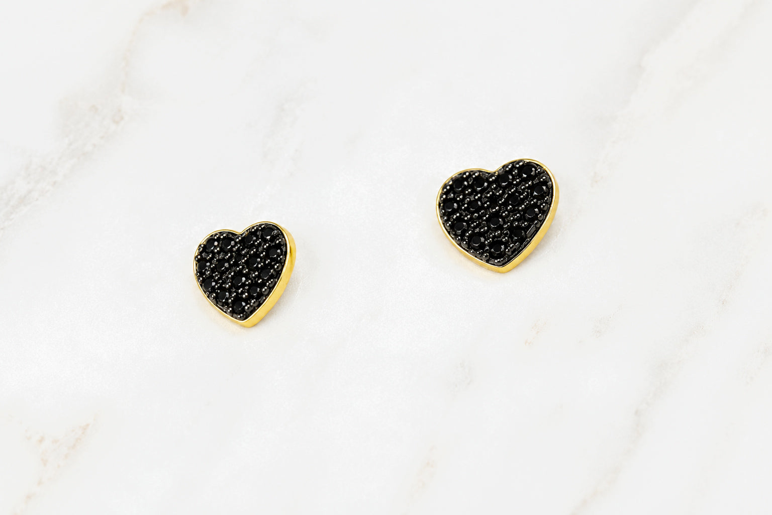 A pair of small heart-shaped stud earrings with textured black pavé centers and polished gold borders, displayed on a white marble surface.