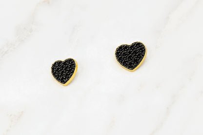 A pair of small heart-shaped stud earrings with textured black pavé centers and polished gold borders, displayed on a white marble surface.
